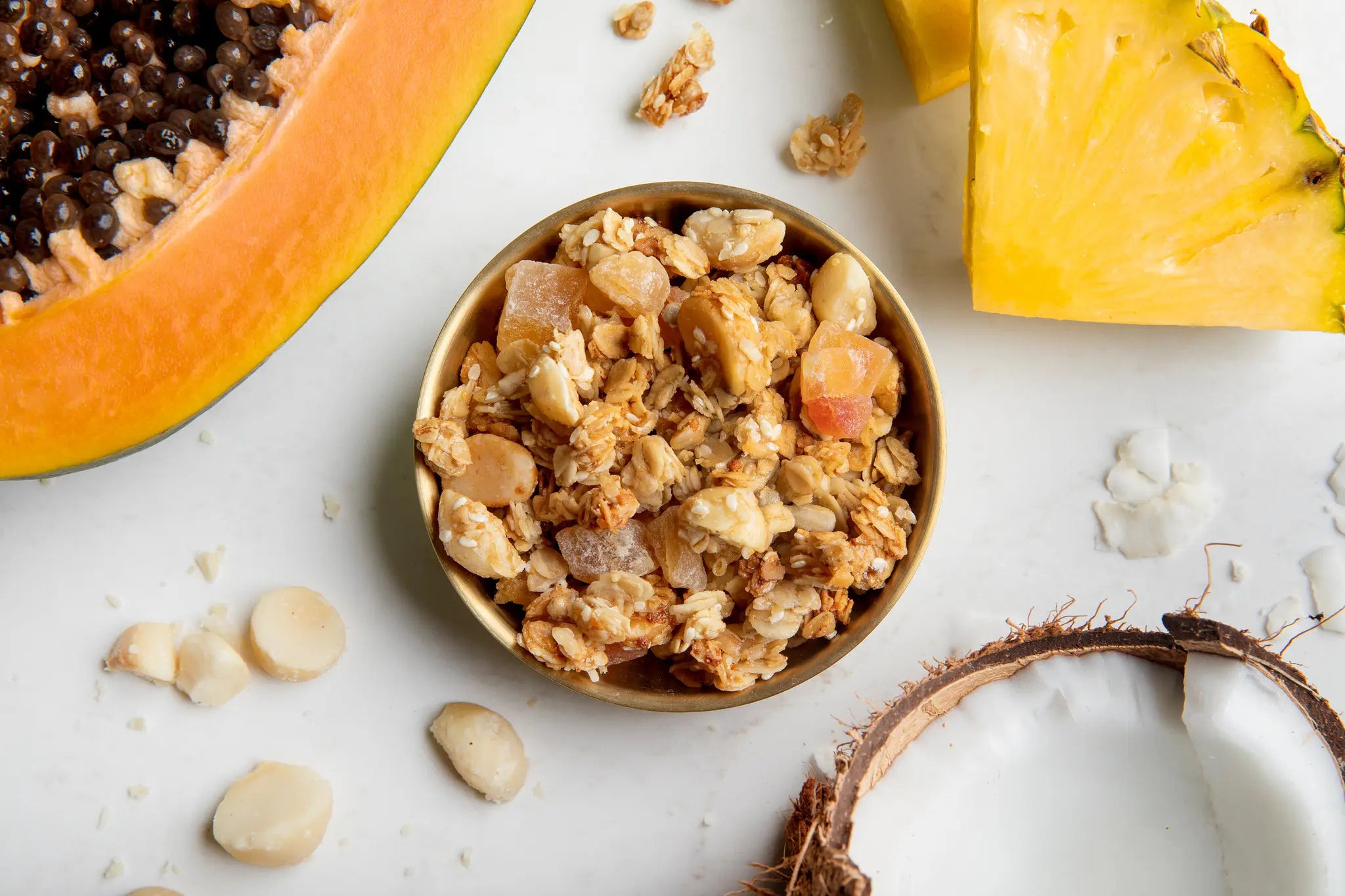 Small bowl of granola with a halved papaya, pineapple, and coconut on a granite countertop